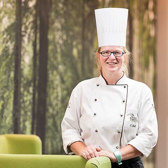 Blonde Köchin mit Kochmütze und Kochjacke steht an einem apfelgrünem Sofa und lächelt in die Kamera.