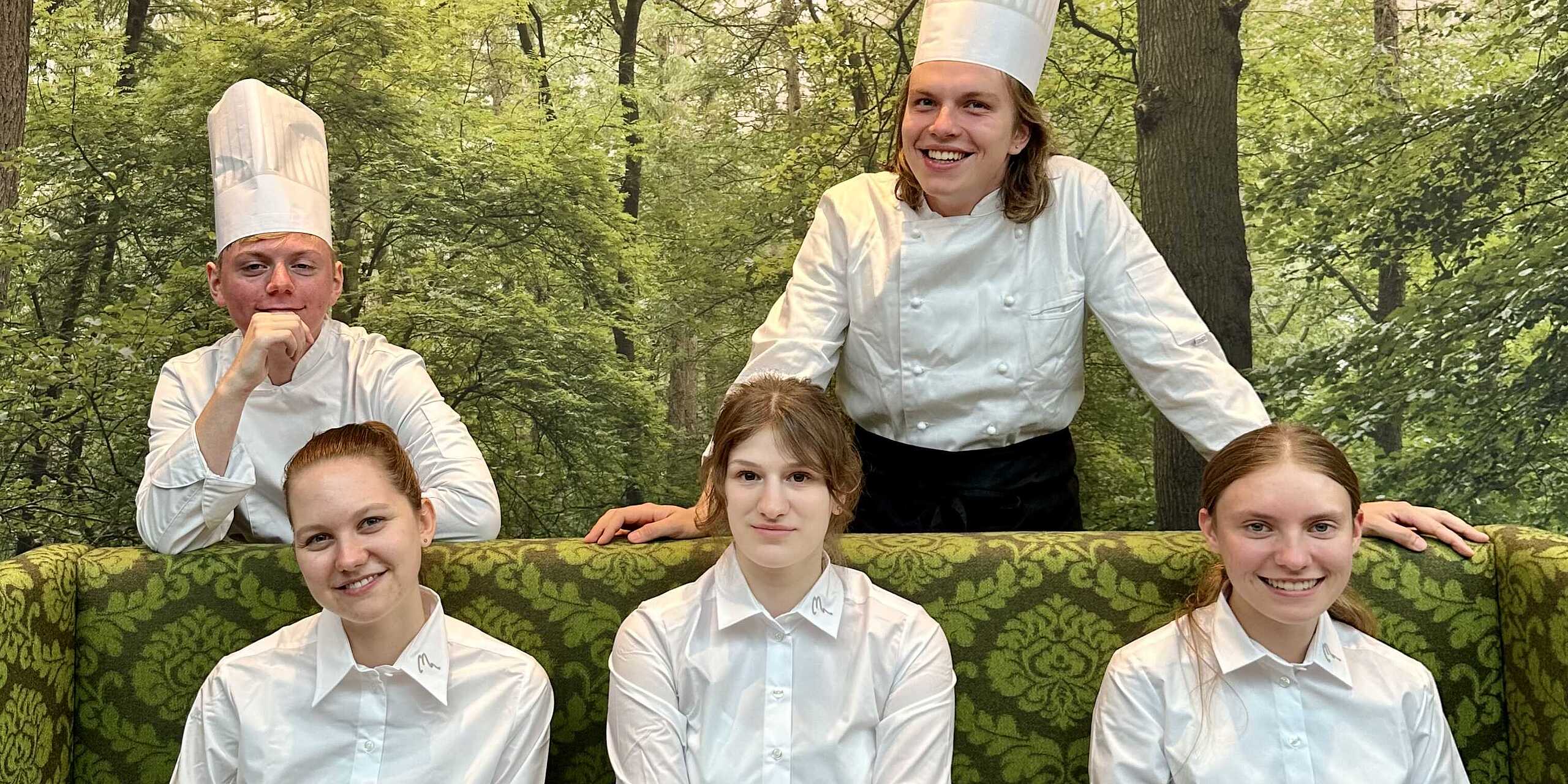 zwei männliche Kochauszubildende in Kochbekleidung und drei junge Mädchen in weißen Uniformhemden  
