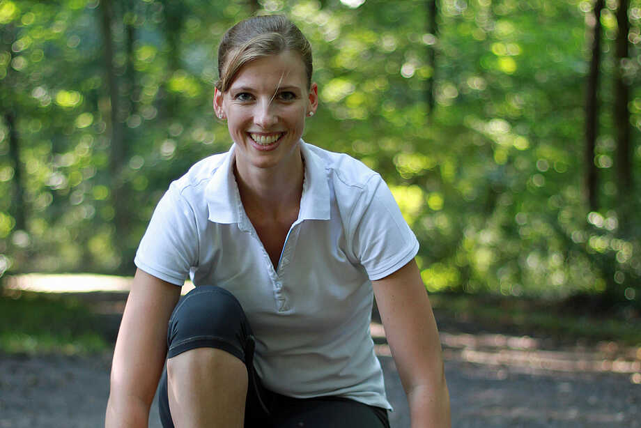 Sina Cordsen - Frau mit weißem T-Shirt guckt im Wald lächelnd geradeaus in die Kamera