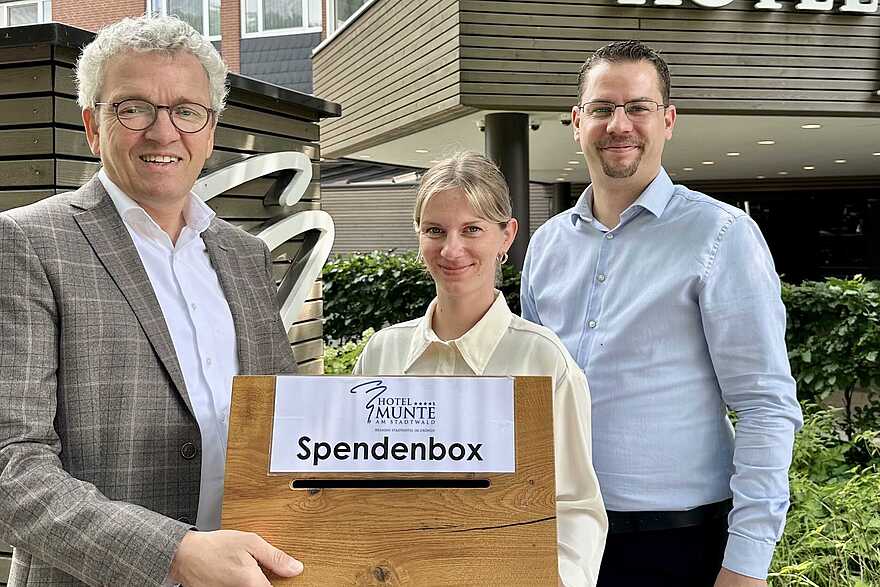 zwei vor einem Hotel stehende Männer und eine Frau lächeln in die Kamera