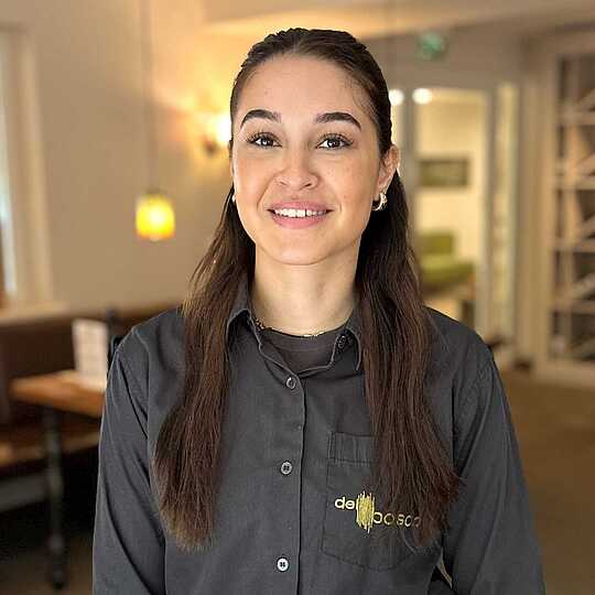 Auszubildende in dunkler Dienstkleidung mit Logo der Trattoria "del bosco" blickt freundlich in die Kamera. Im Hintergrund ist ein stilvoll eingerichteter Gastronomiebereich zu sehen.