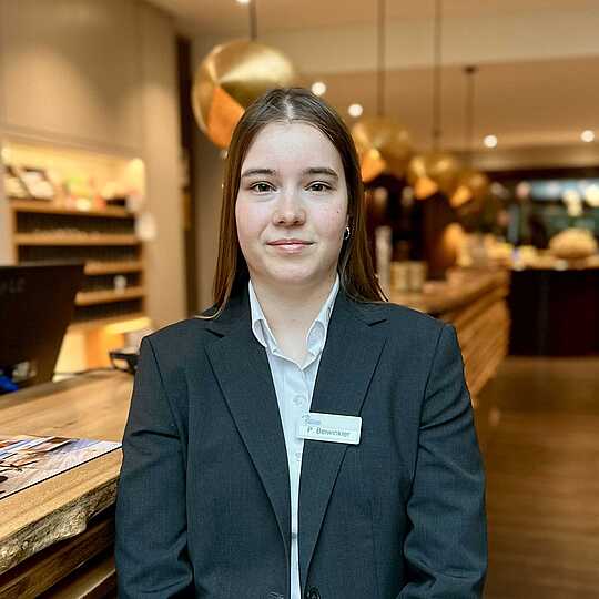 Mitarbeiterin in dunklem Blazer und weißer Bluse steht vor der Rezeption des Hotels.
