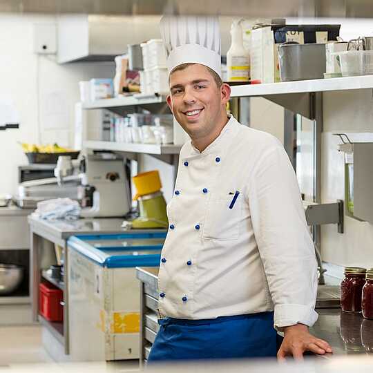 Koch mit Kochmütze in weißer Kochjacke und blauer Schürze steht in einer Großküche und lächelt in die Kamera