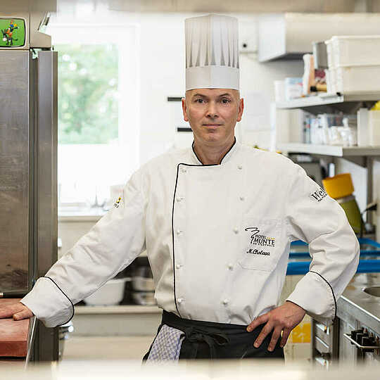 Koch mit Kochmütze steht in stolzer Pose in einer Restaurantküche und lächelt verschmitzt in die Kamera