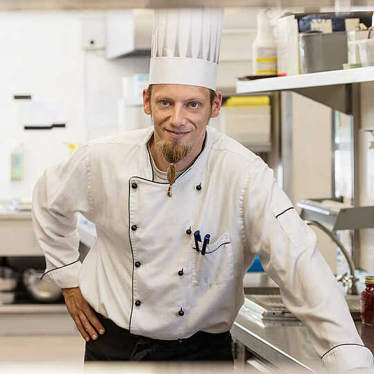 Blonder Koch mit Ziegenbart und Kochmütze steht in einer Küche mit Salzschütten und gefüllten Marmeladengläsern und lächelt in die Kamrera.