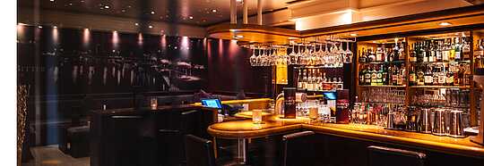 Eine Bar mit beleuchtetem Tresen, vielen Flaschen und Gläsern im Regal sowie mehreren Barhockern.