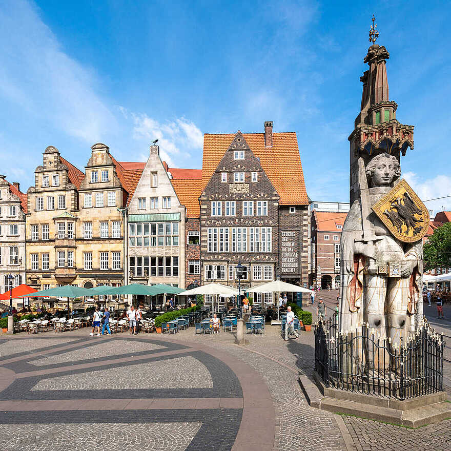 Eine große Statue mit Schild steht auf einem gepflasterten Platz vor historischen Gebäuden mit roten Dächern und Straßencafés.