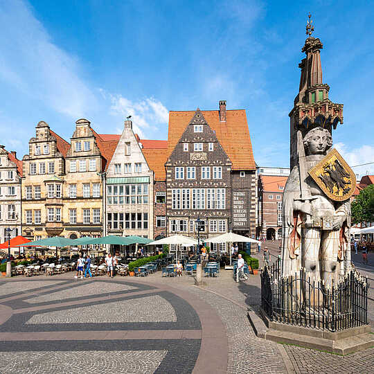 Eine große Statue mit Schild steht auf einem gepflasterten Platz vor historischen Gebäuden mit roten Dächern und Straßencafés.